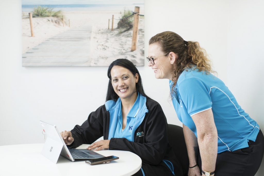 A woman smiles at the camera while she shows another therapist information on a tablet