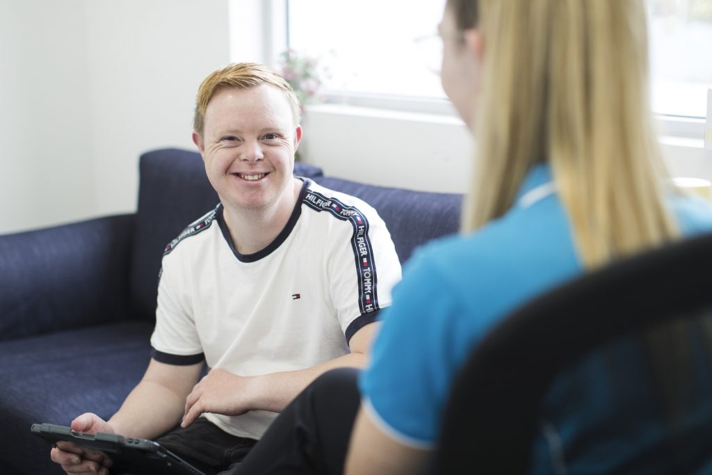 A young man smiles while holding a tablet, facing a therapist